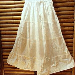 May include: A white tiered skirt with eyelet detailing and a drawstring waist. The skirt is hanging on a hanger against a bamboo background. The skirt is made of a lightweight fabric.