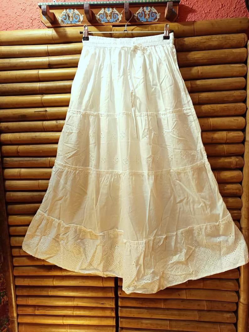 May include: A white tiered skirt with eyelet detailing and a drawstring waist. The skirt is hanging on a hanger against a bamboo background. The skirt is made of a lightweight fabric.