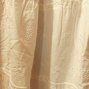 May include: Close-up of a white fabric with intricate eyelet embroidery. The fabric has a textured appearance with a layered design, featuring repeating floral patterns. The overall aesthetic is soft and delicate, suggesting a garment or home decor item.
