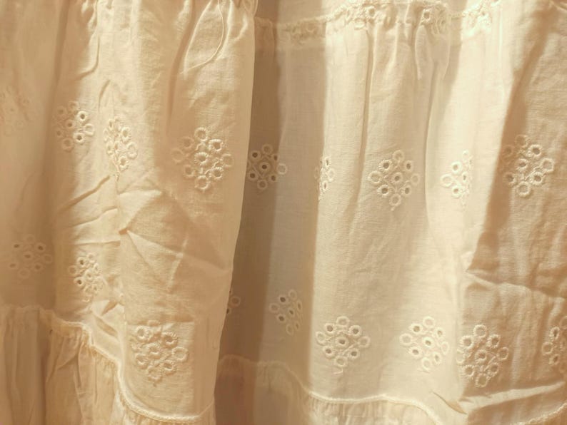 May include: Close-up of a white fabric with intricate eyelet embroidery. The fabric has a textured appearance with a layered design, featuring repeating floral patterns. The overall aesthetic is soft and delicate, suggesting a garment or home decor item.