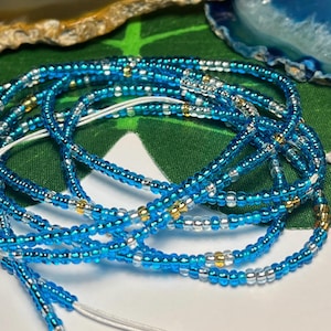 May include: Close-up of several strands of blue, silver, and gold waist beads. The beads are small and strung on a white cord. The beads are arranged in a circular pattern. The background is a green leaf.