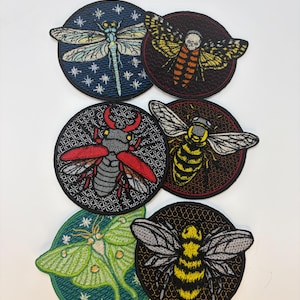 May include: A selection of embroidered insect patches. Designs include a dragonfly, moth with a skull, beetle, bee, and a green moth. Each patch is circular with detailed stitching and vibrant colours.