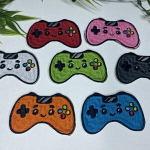 Op de afbeelding: Set van tien geborduurde strijkpatches met een cartoon-videogamecontroller met een glimlachend gezicht. De patches zijn in verschillende kleuren, waaronder rood, roze, groen, grijs, oranje en blauw.