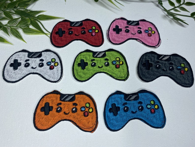Op de afbeelding: Set van tien geborduurde strijkpatches met een cartoon-videogamecontroller met een glimlachend gezicht. De patches zijn in verschillende kleuren, waaronder rood, roze, groen, grijs, oranje en blauw.