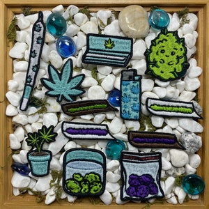 May include: A collection of embroidered patches featuring various cannabis-related imagery, including a joint, a lighter, a bag of weed, and a plant. The patches are arranged on a white background with small white stones.