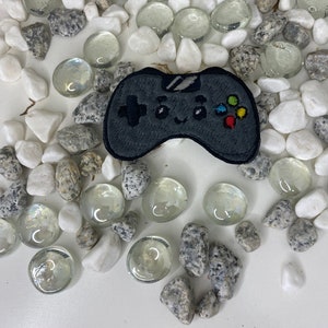Op de afbeelding: Een geborduurde patch van een zwarte en grijze videogamecontroller met kleurrijke knoppen. De patch is omringd door witte en grijze stenen.