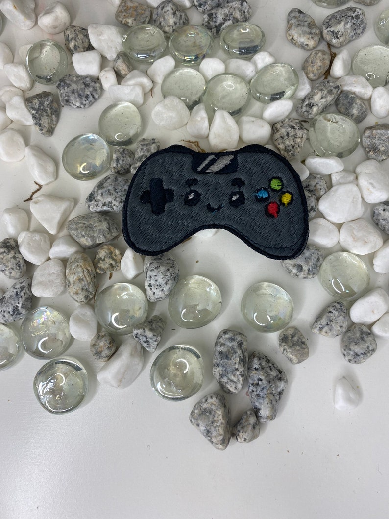 Op de afbeelding: Een geborduurde patch van een zwarte en grijze videogamecontroller met kleurrijke knoppen. De patch is omringd door witte en grijze stenen.