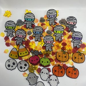 May include: A collection of colourful Halloween-themed iron-on patches featuring mummies, pumpkins, candy corn, ghosts, and apples.