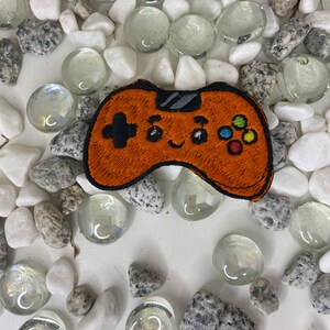 Op de afbeelding: Oranje geborduurde videogamecontroller-patch met een glimlachend gezicht en kleurrijke knoppen. De patch is omringd door witte en grijze stenen.