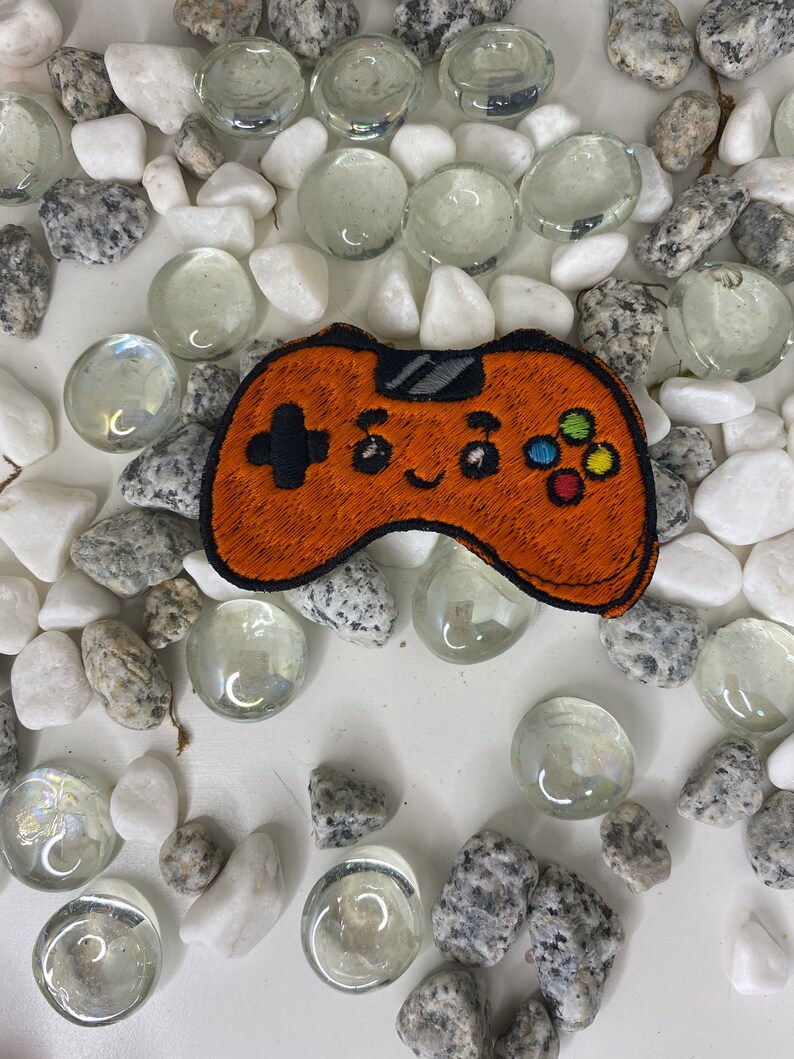 Op de afbeelding: Oranje geborduurde videogamecontroller-patch met een glimlachend gezicht en kleurrijke knoppen. De patch is omringd door witte en grijze stenen.