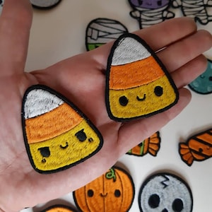 Może przedstawiać: Zbiór naszywek na Halloween z motywem ducha, mumii, kukurydzy cukrowej, dyni i czaszki. Naszywki są haftowane kolorowymi detalami i uroczymi buziami.