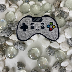 Op de afbeelding: Een grijze en witte geborduurde patch van een videogamecontroller met een glimlachend gezicht. De controller heeft een zwarte d-pad, een zwarte A-knop en een kleurrijke knopset.