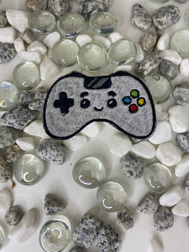 Op de afbeelding: Een grijze en witte geborduurde patch van een videogamecontroller met een glimlachend gezicht. De controller heeft een zwarte d-pad, een zwarte A-knop en een kleurrijke knopset.