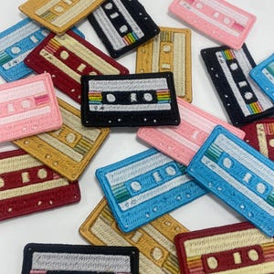 May include: A collection of colorful embroidered cassette tape patches. The patches are in various colors, including red, blue, yellow, pink, and black. Each patch features a white cassette tape with a rainbow-colored tape.