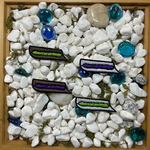 May include: Four embroidered patches with a sandwich design. The patches are white with green, purple, and brown embroidery. The patches are on a white background with small white rocks.