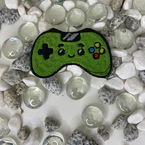 Op de afbeelding: Groene geborduurde videogamecontroller-patch met een glimlachend gezicht en kleurrijke knoppen. De patch is omringd door witte en grijze stenen.