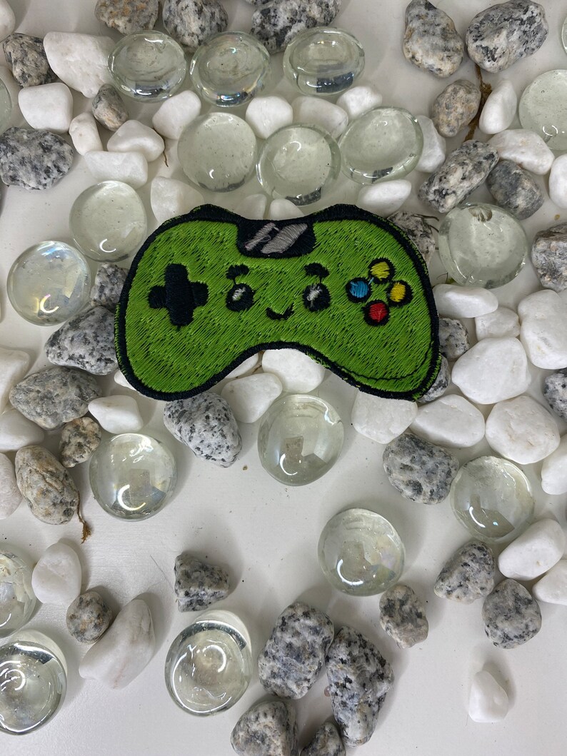 Op de afbeelding: Groene geborduurde videogamecontroller-patch met een glimlachend gezicht en kleurrijke knoppen. De patch is omringd door witte en grijze stenen.