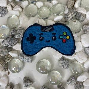 Op de afbeelding: Een blauwe geborduurde videogamecontroller-patch met kleurrijke knoppen. De patch heeft een glimlachend gezicht en is omringd door witte en grijze stenen.