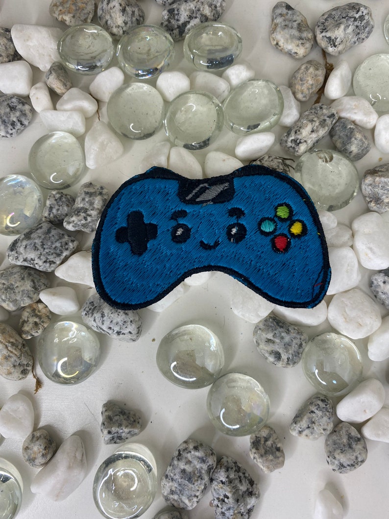 Op de afbeelding: Een blauwe geborduurde videogamecontroller-patch met kleurrijke knoppen. De patch heeft een glimlachend gezicht en is omringd door witte en grijze stenen.