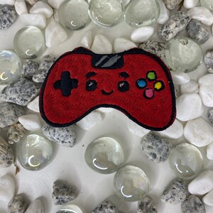 Op de afbeelding: Rode geborduurde videogamecontroller-patch met een glimlachend gezicht en kleurrijke knoppen.