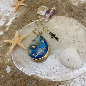 May include: A gold chain necklace with a teardrop-shaped pendant filled with blue resin, glitter, and seashells. The pendant is surrounded by a gold frame. A small gold key charm hangs from the chain.