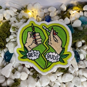 Può includere: Una toppa ricamata verde e gialla a forma di cuore spezzato. Il lato sinistro del cuore presenta una mano che tiene un accendino e il testo "BEST". Il lato destro del cuore presenta una mano che tiene uno spinello e il testo "BUDS".