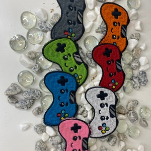 Puede incluir: Siete parches bordados de mando de juego en diferentes colores: gris, naranja, rojo, verde, azul, blanco y rosa. Los parches están dispuestos en semicírculo sobre un fondo blanco con piedras blancas.