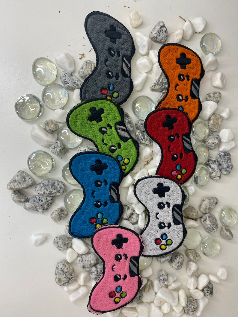 Op de afbeelding: Zeven geborduurde gamecontroller-patches in verschillende kleuren: grijs, oranje, rood, groen, blauw, wit en roze. De patches zijn in een halve cirkel op een witte achtergrond met witte stenen gerangschikt.