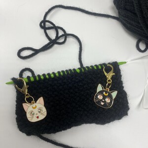 May include: Black knitting project with green yarn and two gold cat charms attached. One charm is a white cat with a crescent moon and the other is a black cat with a crescent moon.