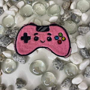 Op de afbeelding: Een roze geborduurde videogamecontroller-patch met een schattig gezicht en kleurrijke knoppen. De patch is omringd door witte en grijze stenen.