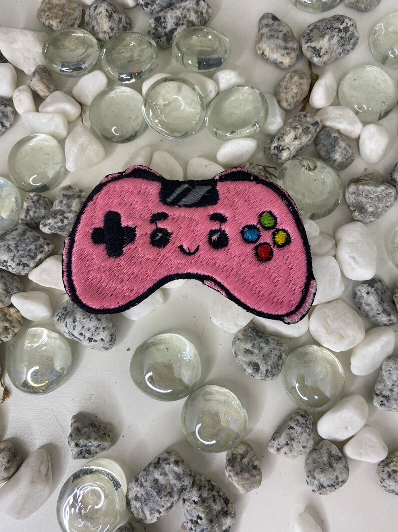Op de afbeelding: Een roze geborduurde videogamecontroller-patch met een schattig gezicht en kleurrijke knoppen. De patch is omringd door witte en grijze stenen.