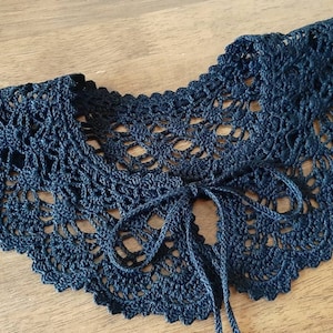 May include: A black crochet collar featuring an intricate, openwork lace pattern with a scalloped edge. The accessory includes two thin, matching black crochet ties at the center front, designed to be fastened in a bow for a secure fit around the neck.