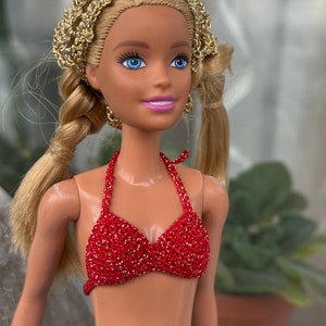 May include: A blonde doll wearing a red crocheted bikini top with gold accents. The doll has blue eyes and is wearing a gold headband.