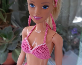May include: A doll with blonde hair wearing a pink crocheted bikini and a matching pink crocheted headscarf. The bikini top and bottom are decorated with small white beads along the edges, and the bikini bottom features side ties.