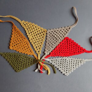 May include: A collection of six triangular crochet bandanas featuring an openwork lattice pattern. The set includes individual pieces in olive green, mustard yellow, gold, beige, bright red, and light gray, each with thin ties at the corners.