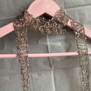May include: A long, narrow scarf made of a sheer, open-knit metallic brown yarn. The fabric is densely embellished with small, reflective sequins that catch the light, creating a shimmering effect throughout the entire length of the accessory.