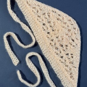 May include: A light beige knitted headband with a delicate openwork design. The headband has long, thin ties on each side for adjusting the fit. The soft, textured material is visible against a dark blue background.