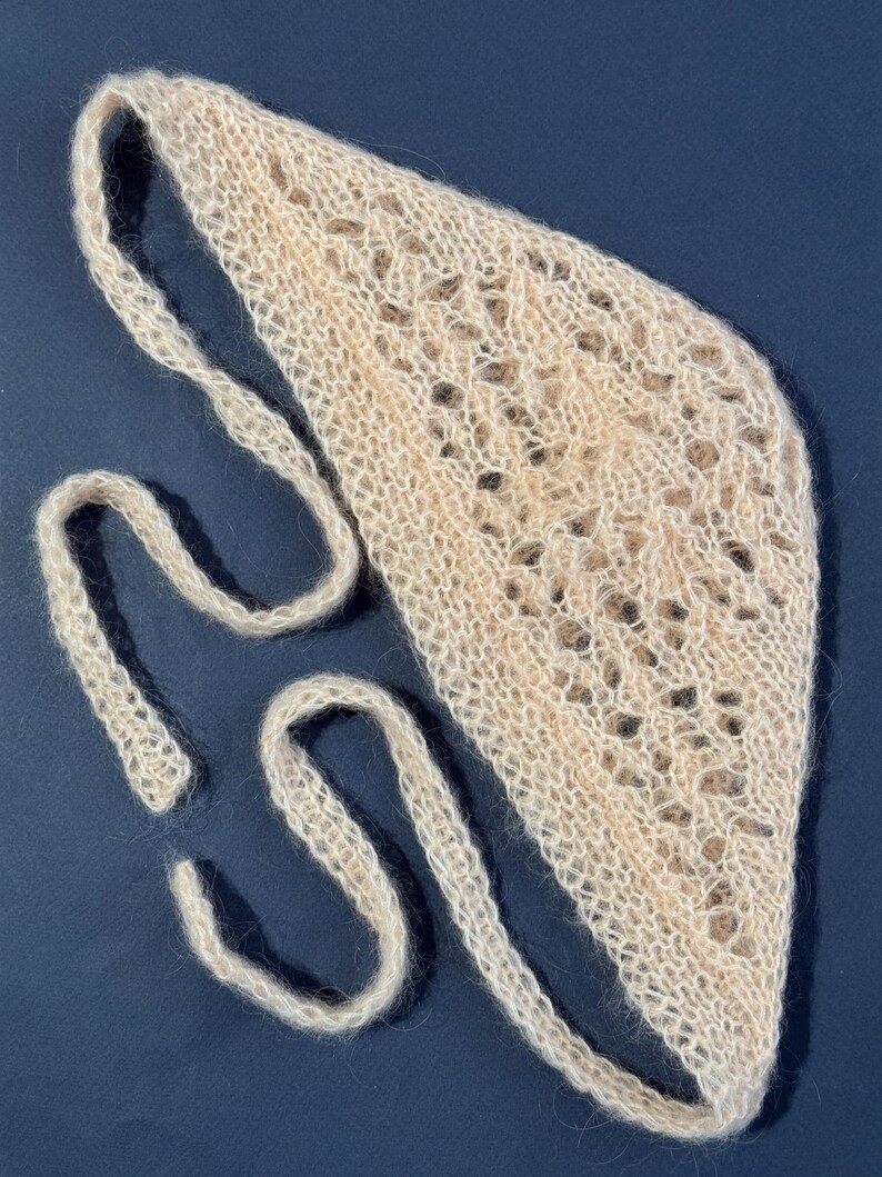 May include: A light beige knitted headband with a delicate openwork design. The headband has long, thin ties on each side for adjusting the fit. The soft, textured material is visible against a dark blue background.