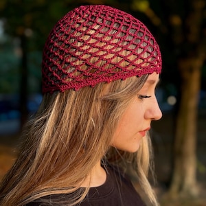 May include: A burgundy crochet beanie with a delicate open-weave pattern. The beanie is worn on a person with long blonde hair.