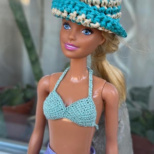 May include: A doll wearing a blue and white crocheted hat, a light blue crocheted bikini top, and a lavender skirt.