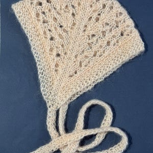 May include: A cream-colored, hand-knitted baby bonnet with a delicate openwork pattern. The bonnet features a fan-shaped crown and long ties for securing. The soft, textured yarn creates a cozy and vintage aesthetic.