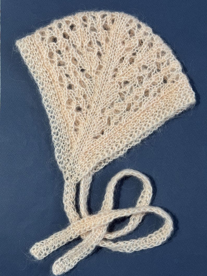 May include: A cream-colored, hand-knitted baby bonnet with a delicate openwork pattern. The bonnet features a fan-shaped crown and long ties for securing. The soft, textured yarn creates a cozy and vintage aesthetic.