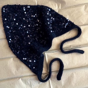 May include: A black knitted hat with sequins and long ties. The hat is triangular in shape and has a sparkly finish.