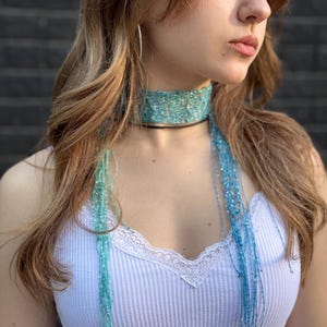 May include: A turquoise and light blue crocheted choker necklace featuring a wide, textured band that wraps around the neck, with long, thin, dangling strands of matching yarn extending downwards from the centre of the piece.