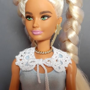 May include: A blonde doll with green eyes wearing a white crocheted collar and a light blue sleeveless top. The doll is wearing gold star-shaped earrings and a gold chain necklace.