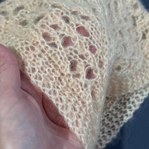 May include: A delicate, open-weave bonnet in a soft, pale peach color. The knitted fabric has a lacy pattern with small holes throughout. The bonnet is held in a hand, showcasing the texture and fine detail of the yarn. The style is vintage and handmade.
