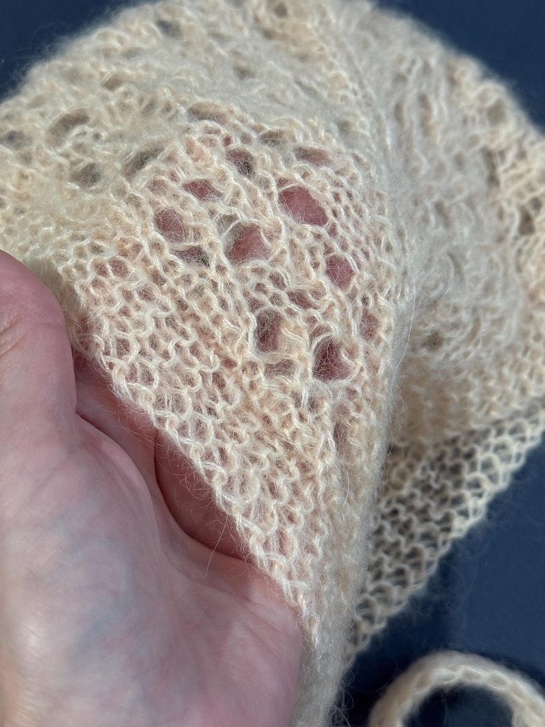 May include: A delicate, open-weave bonnet in a soft, pale peach color. The knitted fabric has a lacy pattern with small holes throughout. The bonnet is held in a hand, showcasing the texture and fine detail of the yarn. The style is vintage and handmade.