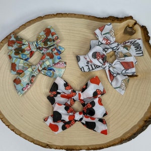 May include: Three fabric hair bows with different patterns are displayed on a wooden slice. One bow features a pattern of gingerbread men and candy canes on a light blue background. Another has a Minnie Mouse pattern with red bows and black polka dots. The third bow has a pattern of various objects.