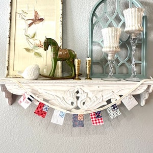 May include: A white shelf with decorative carvings holds a framed print of a bird, a green wooden rocking horse, two candle holders, and a white bird figurine. A fabric banner with red, white, and blue patterned flags hangs below the shelf.