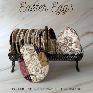 Puede incluir: Huevos de Pascua de tela reutilizables en un soporte de metal decorativo. Los huevos presentan varios diseños, incluyendo florales, de conejos y toile de Jouy. El soporte contiene varios huevos, con un huevo en primer plano. El texto dice "Reusable Fabric Easter Eggs".